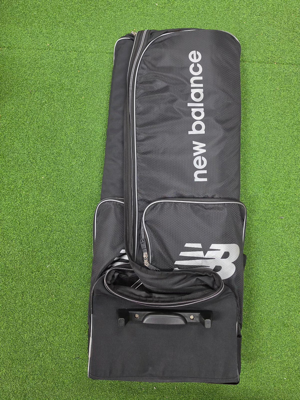 Top-down flat view of a premium black New Balance 10i combo duffle wheelie cricket kit bag on green artificial grass.