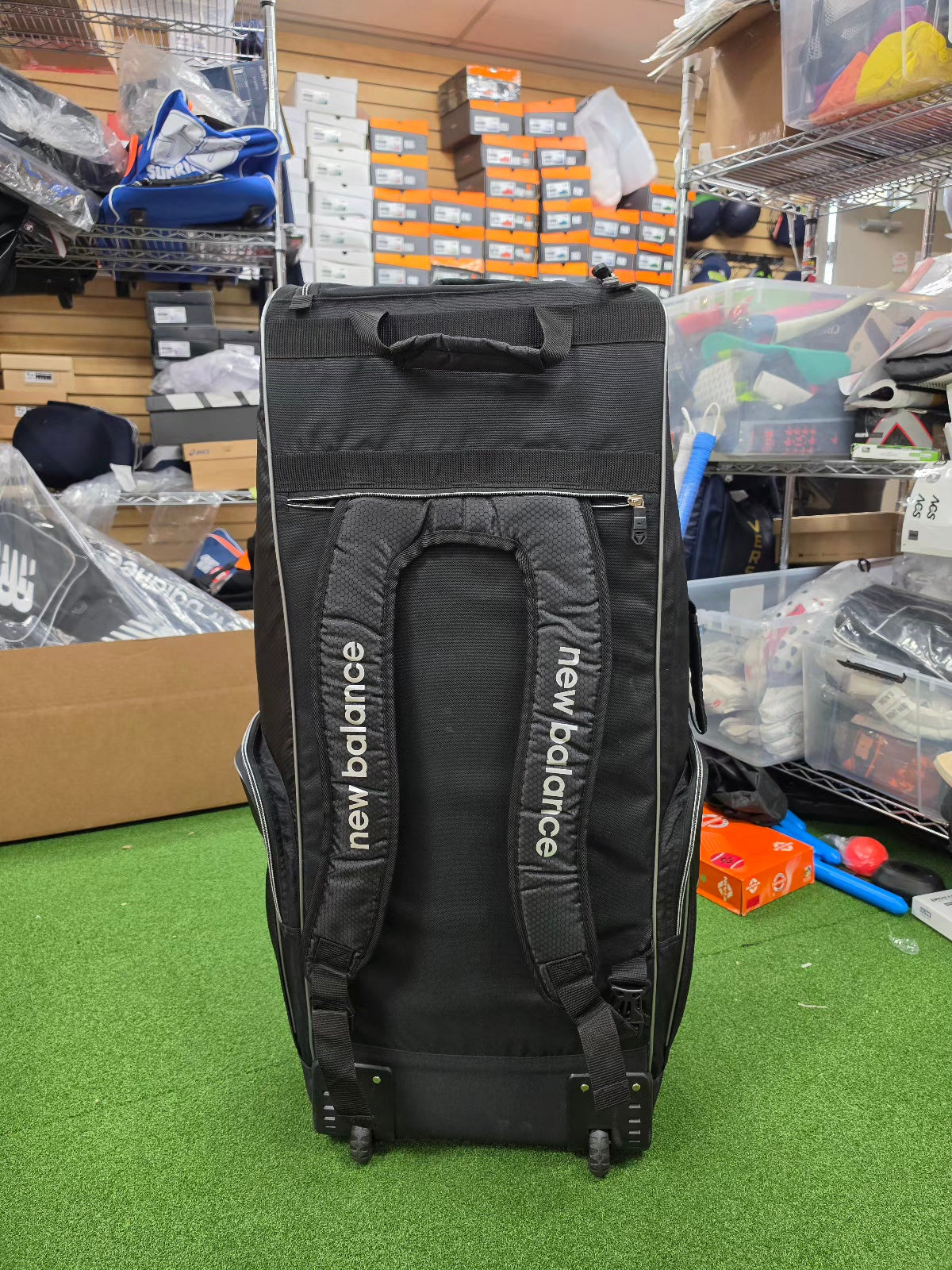Rear view of black New Balance 10i combo duffle wheelie cricket kit bag showing padded backpack shoulder straps standing in a shop.