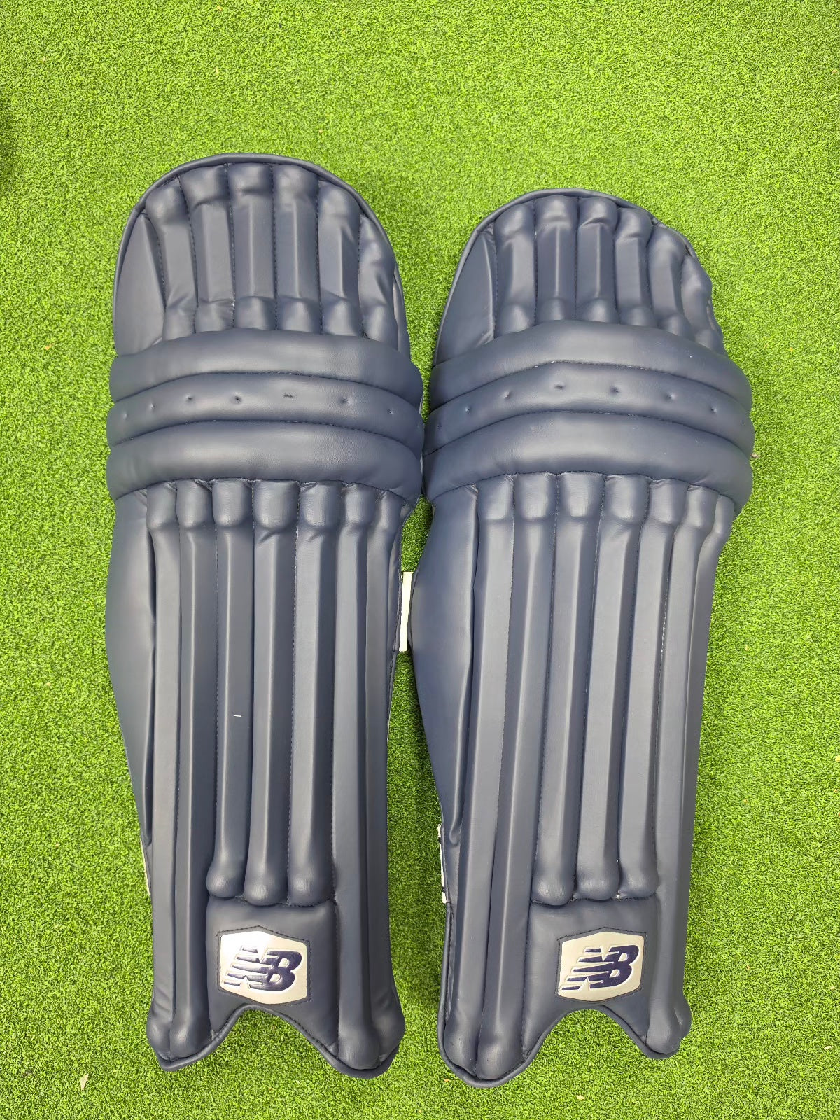 Pair of cricket batting pads fitted with navy blue colored protective clads or covers on a green artificial turf background.