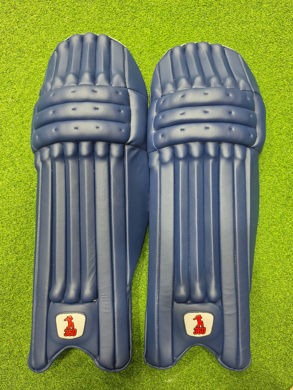Top face view of EM 360 Bold adult navy blue cricket batting pads on green artificial turf on Alberta.