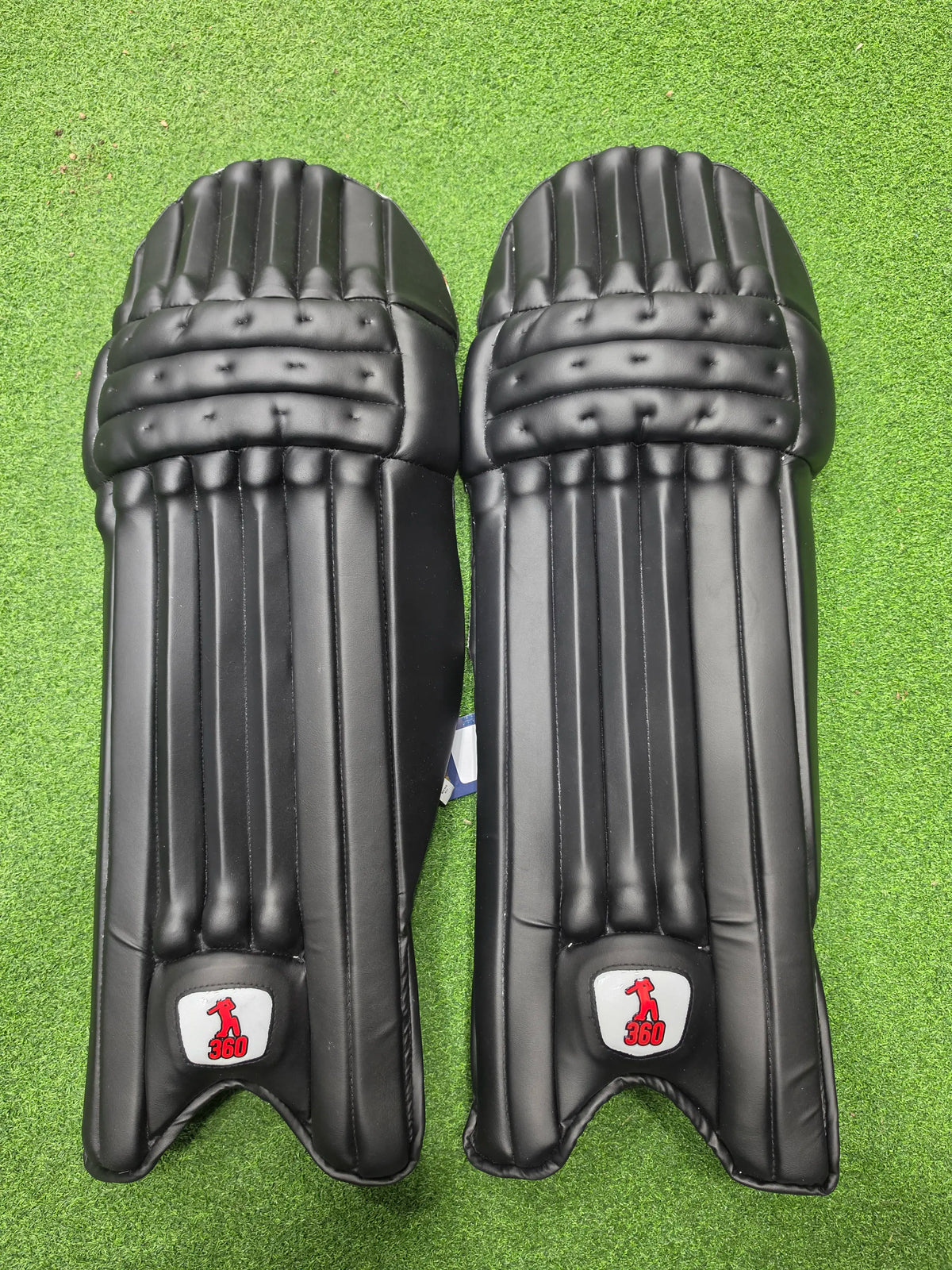 Front face and inner padded back view of EM 360 Bold adult black cricket batting pads with red accents on artificial grass on Alberta.
