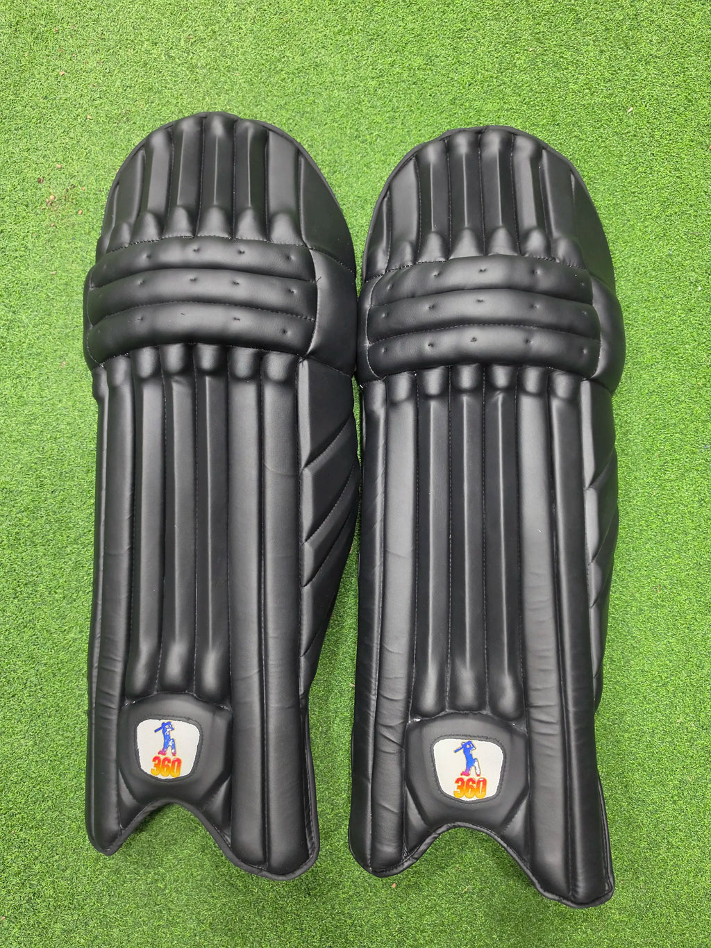 Top face view of EM 360 PLAY BURST adult white cricket batting pads on green artificial turf on Alberta.