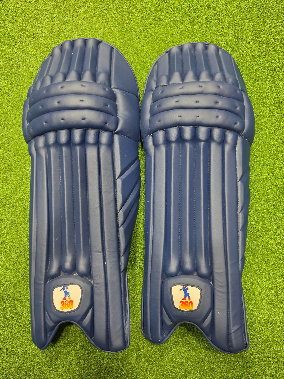 Top face view of EM 360 PLAY BURST adult navy blue cricket batting pads on green artificial turf on Alberta.