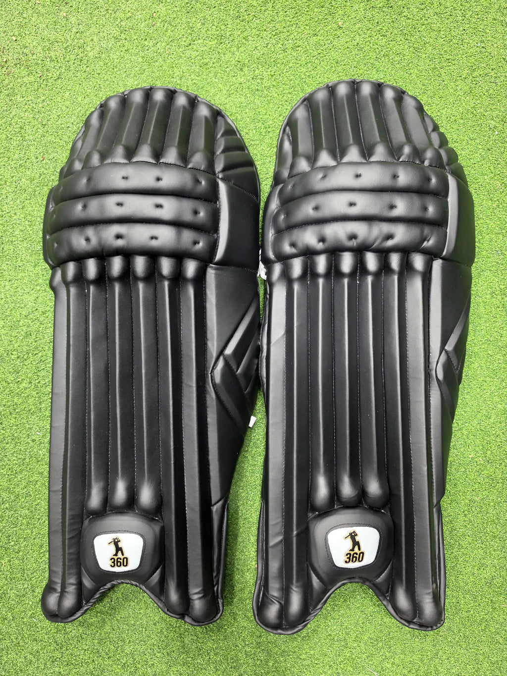 Top view of EM 360 No 17 adult black cricket batting pads on green artificial turf on Alberta.