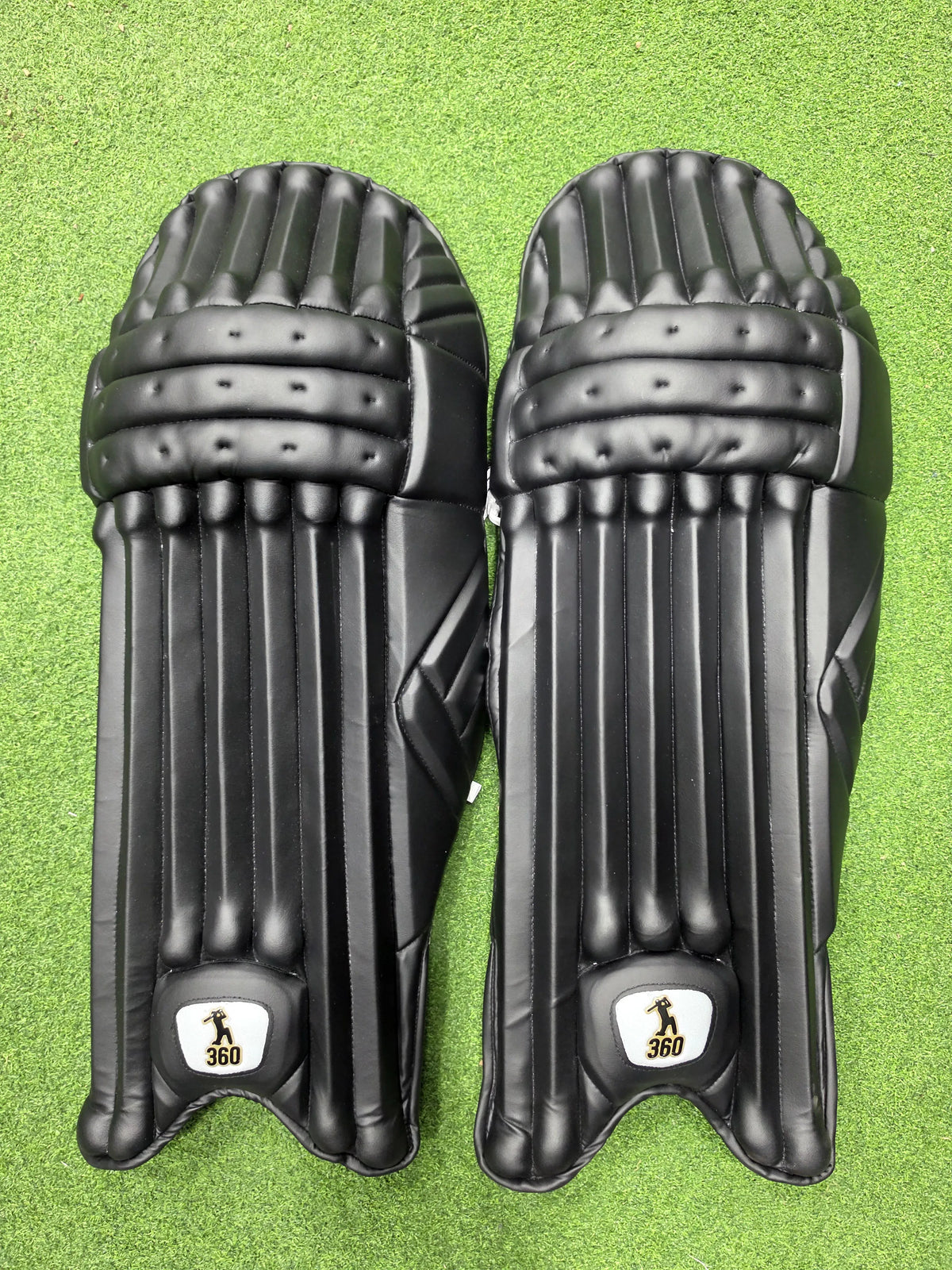 Top view of EM 360 No 17 adult black cricket batting pads on green artificial turf on Alberta.