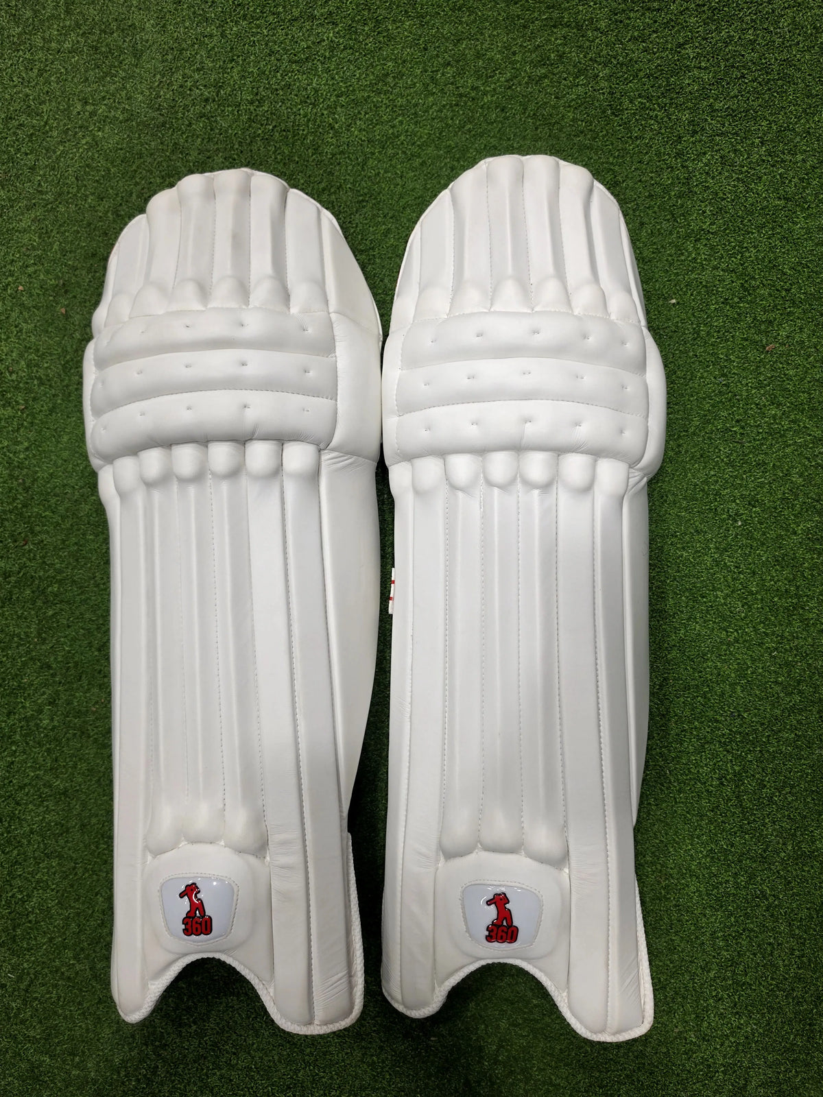 Front face and inner padded back view of EM 360 Bold adult white cricket batting pads with red accents on artificial grass on Alberta.