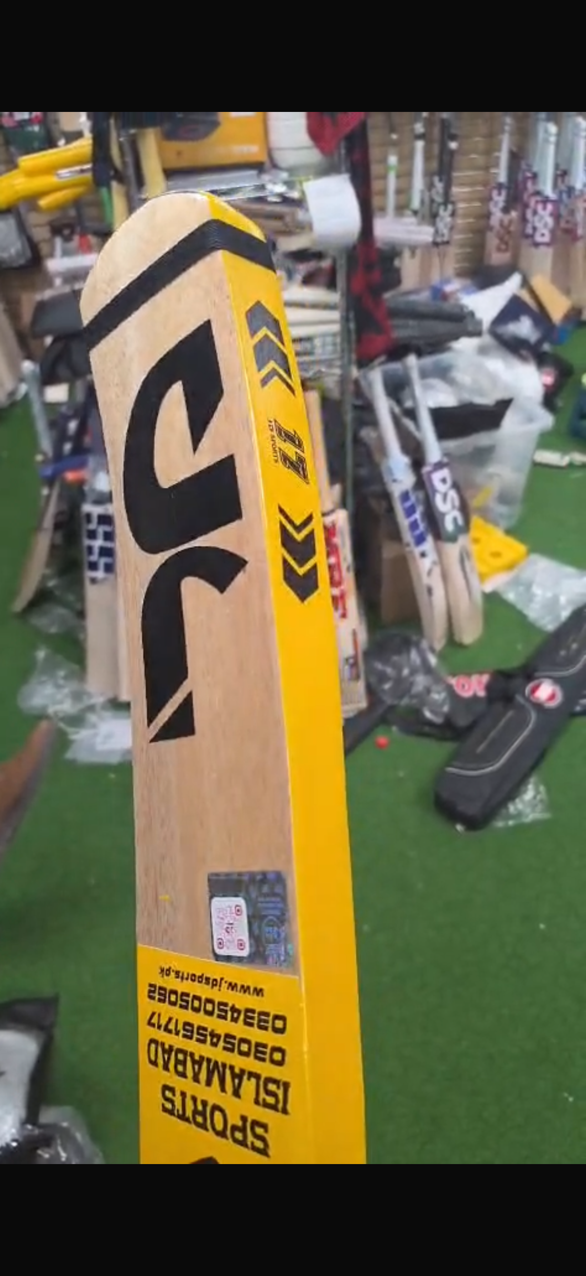 Close-up of a DSC cricket bat with yellow and black branding stickers on a green turf floor background from Albertacricketstore.com.