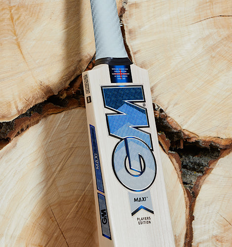 Vertical close-up of the GM Jacob Bethell Players Edition cricket bat face, showcasing the large blue GM stickers and premium handle grip against a background of natural willow tree logs