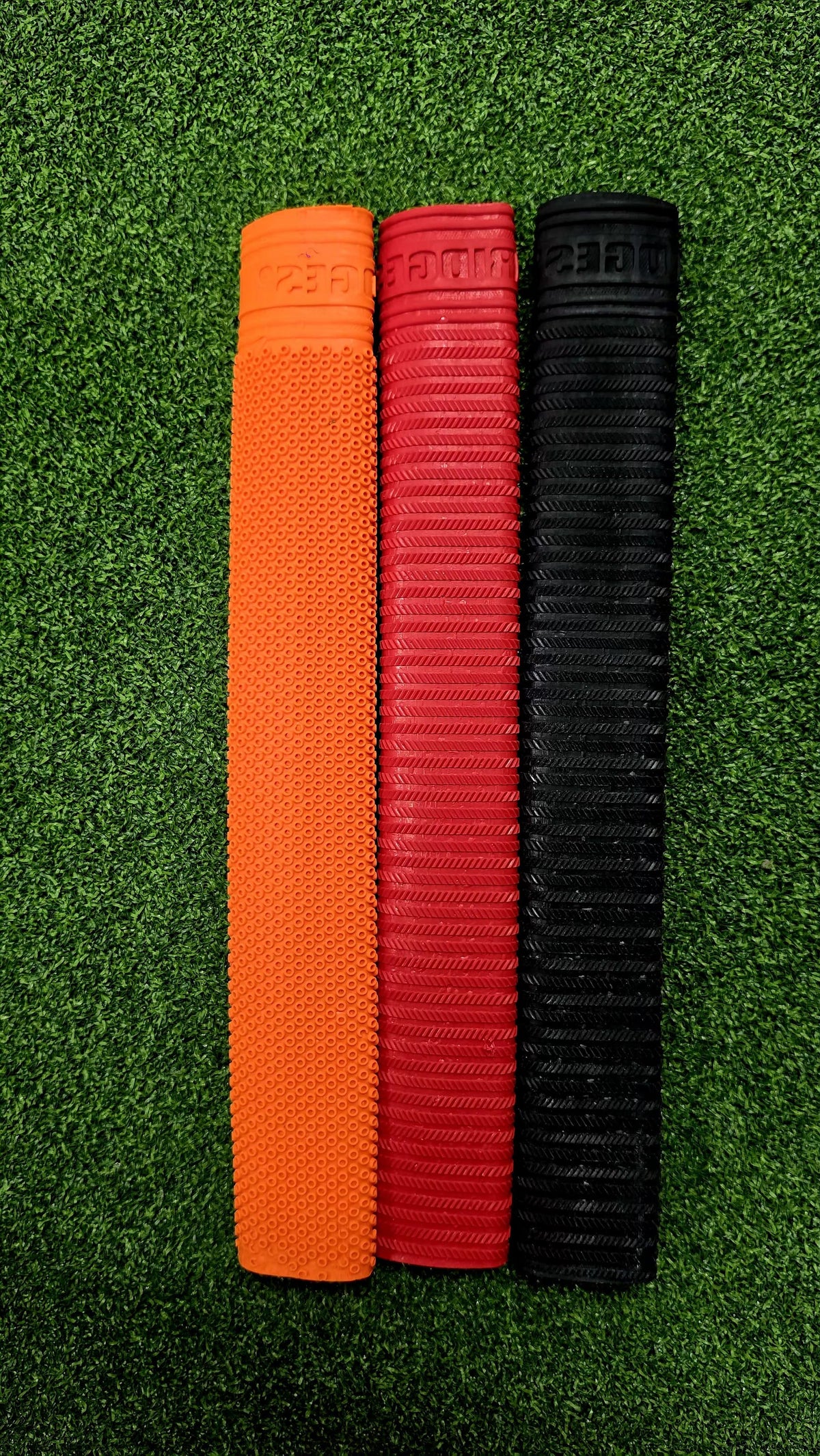Orange, red, and black textured cricket bat grips placed together on a green turf background.